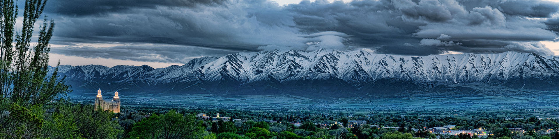 Homepage - Aerial View of Buildings Surrounded by Green Foliage with Snow Covered Mountains in the Background with Dark Clouds