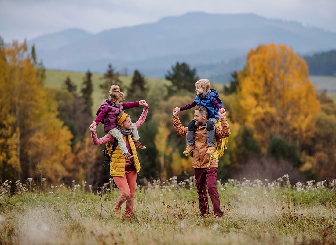 Personal Insurance - View of Two Parents Holding Up Their Children on Their Shoulders During a Hiking Trip on a Field with Flowers with Mountains in the Background